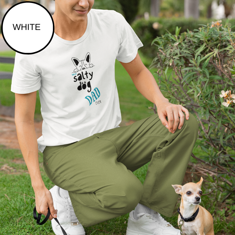 White t-shirt featuring the text 'Best Salty Dog Dad Ever' in playful fonts. The design includes an illustration of a cute dog's face peeking over the words, combining nautical and pet-loving themes. The background showcases a serene sailing scene with ropes and a yacht deck.