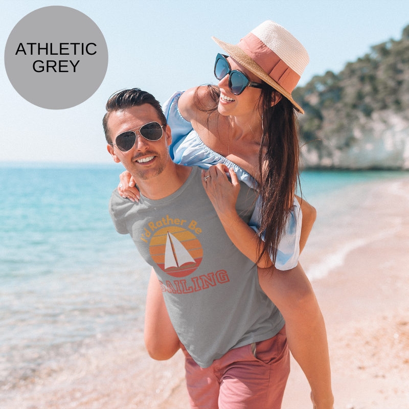 Heather gray t-shirt with the text 'I'd Rather Be Sailing' displayed above and below a retro-style sunset graphic featuring a white sailboat. The vibrant orange, red, and yellow hues of the design contrast with the dark fabric, creating a bold and nautical-themed look. Background shows a peaceful sailing scene with a yacht and open ocean.