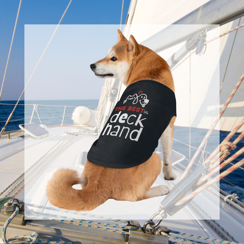 A Shiba Inu dog wearing a black shirt with the text 'The Best Little Deckhand,' sitting proudly on a sailboat deck against a blue ocean backdrop.
