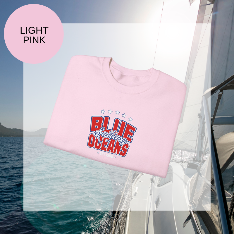 Light pink women's sweatshirt with a bold "Blue Oceans Sailing" print, featuring stars and a nautical-inspired design, displayed against a sailing-themed background.