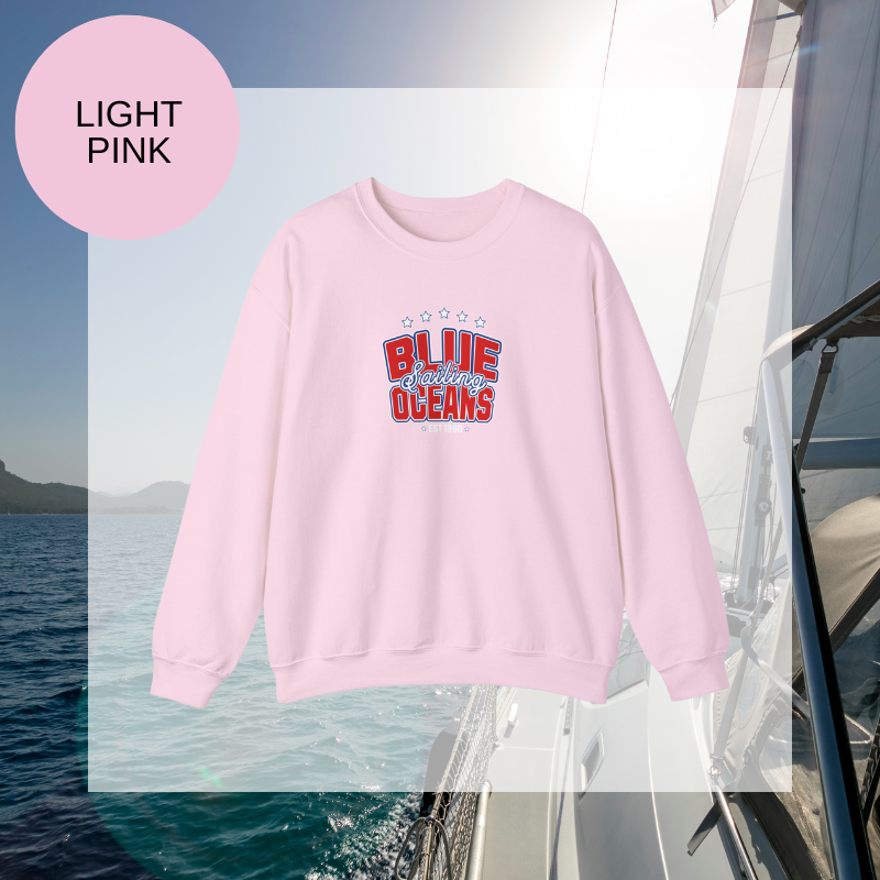 Light pink women's sweatshirt with a bold "Blue Oceans Sailing" print, featuring stars and a nautical-inspired design, displayed against a sailing-themed background.