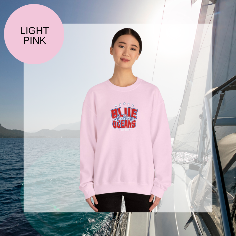 Light pink women's sweatshirt with a bold "Blue Oceans Sailing" print, featuring stars and a nautical-inspired design, displayed against a sailing-themed background.