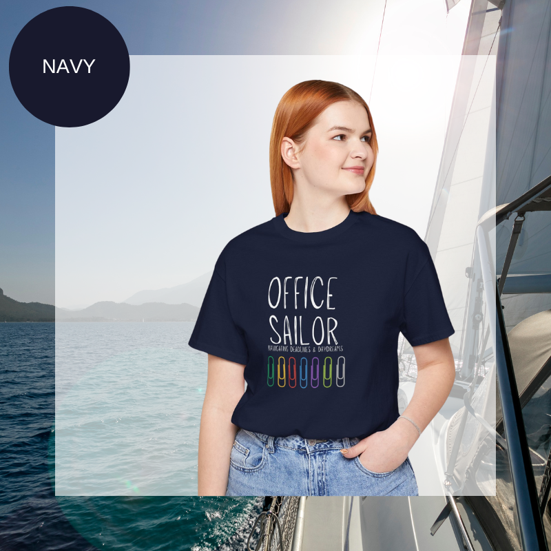 Navy women's t-shirt with the text 'Office Sailor: Navigating Deadlines & Daydreams' and colorful paperclip illustrations, displayed on a sailing-themed background.