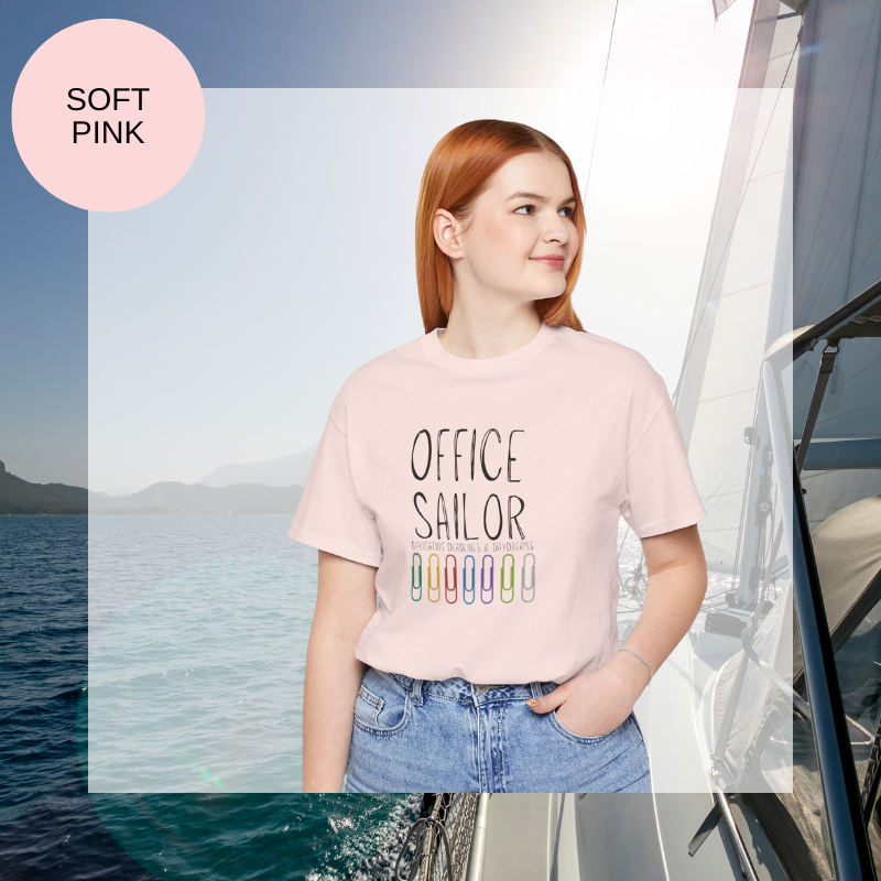 Pink women's t-shirt with the text 'Office Sailor: Navigating Deadlines & Daydreams' and colorful paperclip illustrations, displayed on a sailing-themed background.